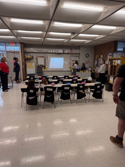 A wide shot of a brightly lit classroom shows a large interactive whiteboard at the front displaying a grid or list of content. An alphabet line with uppercase and lowercase letters A through Z runs across the wall above the main whiteboard. To the left of the main whiteboard, a smaller whiteboard displays simple multiplication equations: "4 x 5 = 20," "2 x 10 = 20," and "5 x 4 = 20." An American flag is mounted above the main whiteboard. An EXIT sign is visible in the classroom. Several adults are present: two women stand on the left side facing the front, two women stand near the main whiteboard in the middle-back area, and another woman stands on the right side. In the immediate foreground on the right, the lower half of a person wearing a dark shirt, khaki shorts, and brown shoes is partially visible. Student desks with black chairs are arranged in rows throughout the classroom, with two children visible seated at desks viewed from behind. The right side of the classroom features a brick wall section and shelving units filled with classroom supplies and containers, some decorated with bee motifs. The classroom has a tiled floor and fluorescent ceiling lights.