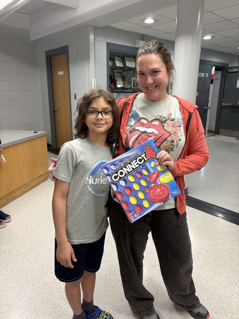 An adult woman and a child stand side-by-side in a institutional building, both smiling at the camera. The woman on the right holds a "Connect 4" board game box labeled "The classic game of CONNECT 4 by Hasbro, Ages 6+." She wears a t-shirt featuring The Rolling Stones tongue logo with floral designs and an orange hooded jacket. The child on the left wears glasses, a grey t-shirt with the "Hurley" brand logo, and navy blue shorts. The background features light grey brick-patterned walls, wooden cabinets, and doors, with one door displaying a "WELCOME TO" sign.