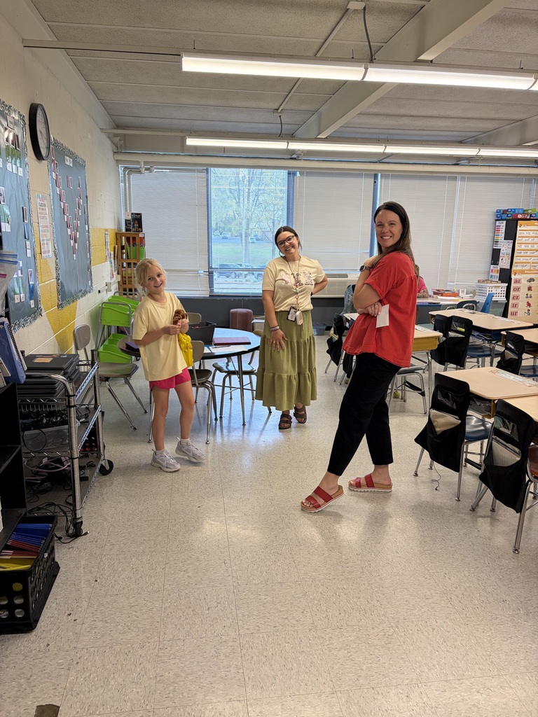 Three females—a young girl and two adult women—stand smiling in a brightly lit classroom. The young girl on the left wears a light yellow t-shirt and pink shorts, holding a small brown plush toy. The woman in the middle wears a light-colored t-shirt with a design, a long olive green skirt, and glasses. The woman on the right wears a red t-shirt and black pants with red sandals, her arms crossed. The classroom features a tiled floor, white walls, and a large window with blinds in the background overlooking trees. Fluorescent lights are visible on the ceiling. On the left wall, a clock and a bulletin board decorated with papers and a yellow border are displayed. A metal shelving unit with electronic devices (possibly laptops) and a black crate with colored folders are visible on the far left. A round table with chairs sits in the middle-left area. On the right side, several student desks and chairs are arranged, some with black storage bags attached to chair backs. A chart on the right wall displays letter combinations "or," "ir," "er," and "ur" repeated multiple times. A blue paper on the left bulletin board shows the partially visible word "CLASS."