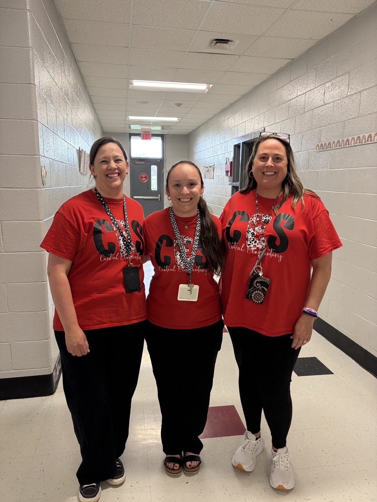 Three women stand side-by-side in a brightly lit school hallway, smiling at the camera. All wear matching red short-sleeved t-shirts with a large black "CES" logo featuring a checkerboard or leopard print pattern, beneath which "Central Elementary" is written. Each woman wears black pants or leggings and has an ID lanyard around her neck. The hallway features white block walls, light-colored tiled flooring, and fluorescent ceiling lighting fixtures. A door with an "EXIT" sign above it is visible in the background.