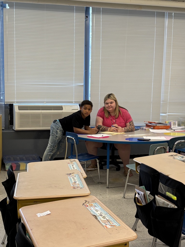 An adult woman with blonde hair and arm tattoos wearing a pink t-shirt sits at a blue oval table with a boy of middle school age wearing a black t-shirt and blue jeans. Both are looking toward the camera. Papers and a pen are on the table in front of the woman. Behind them is a wall with large windows covered by horizontal blinds with ambient light filtering through. A white wall-mounted air conditioning unit is positioned below the windows. In the foreground and to the left are several empty student desks with attached chairs, some with organizing pouches hanging from their sides. The desks feature decorative strips with text and images, with one visible strip reading "LIAM SMARTIN." A box on the table behind the woman is labeled "SIGHT WORDS." The floor is light-colored.