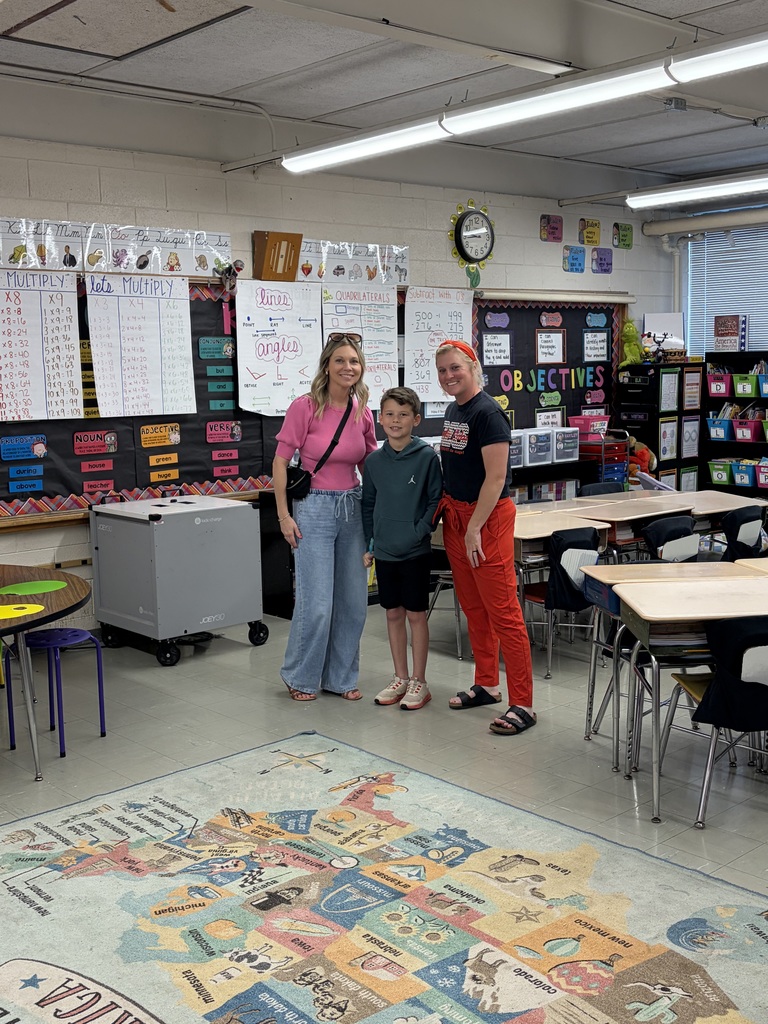 A classroom scene shows three individuals—a woman, a young boy, and another woman—all smiling while standing in the center of the room surrounded by comprehensive educational displays. The walls are adorned with extensive educational charts and posters