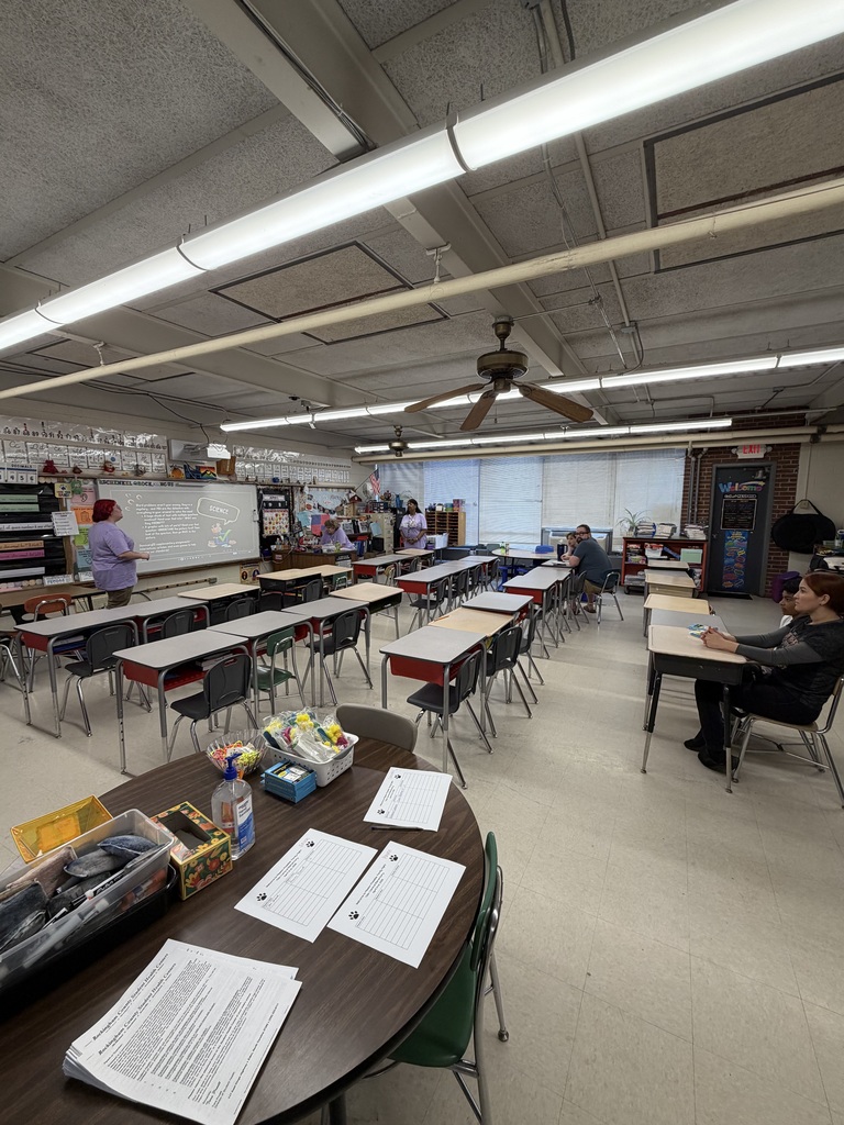 A classroom setting hosts what appears to be a teacher meeting or professional development session. A woman with red hair stands near a projector screen gesturing toward the displayed content. Another woman stands behind a desk in the middle-left, while a man and another person are seated at a table in the middle-right. A woman with dark hair is seated at a desk in the far right foreground. The projector screen displays "SCIENCE topics" with three questions: "How to plan an investigation," "What do we do during an investigation?" and "What happens at the end of an investigation?" Above the screen, wall text reads "SCIENTIFIC PROCESS WE DO." The right wall displays "EXIT" and "WELCOME" signage. Student desks with attached chairs are arranged in rows facing the front. A large round table in the foreground holds papers featuring a paw print logo, a stack of papers labeled "Rockingham County Public Schools Middle School Health Services" with a numbered procedural list, a bottle of hand sanitizer, colorful supplies in baskets and containers, and a black caddy with additional items. The classroom features a light-colored tile floor, a drop ceiling with fluorescent lighting and a ceiling fan, windows with horizontal blinds, and a brick wall section on the right.