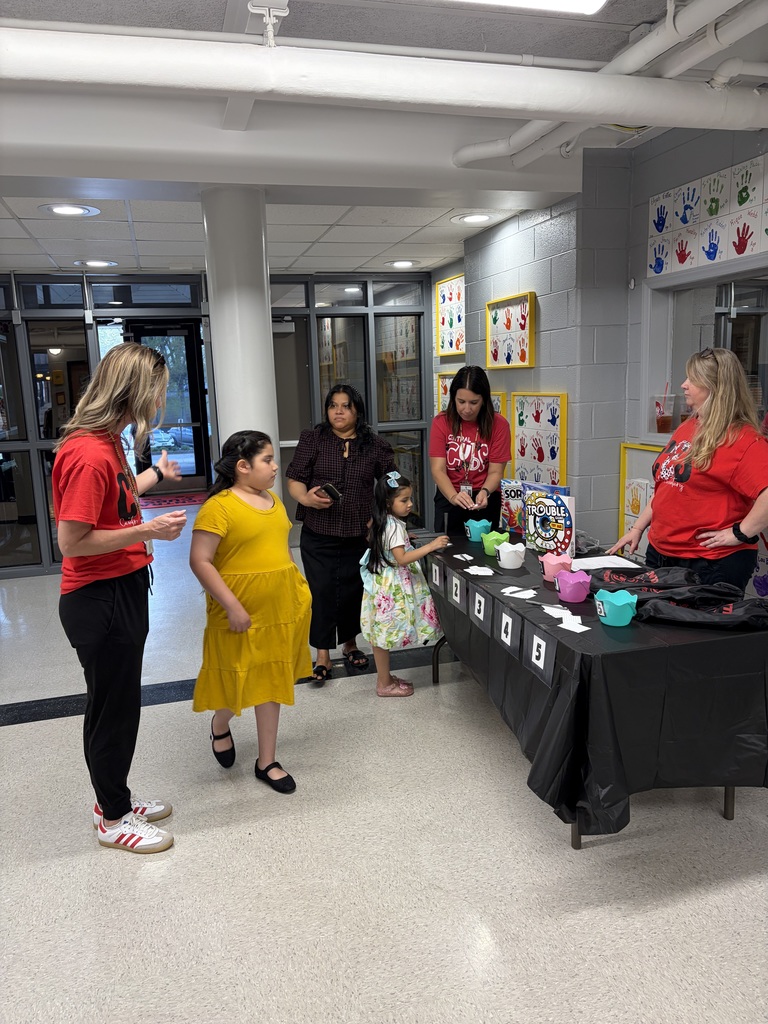 A group of people—primarily women and young girls—gather around a black-draped table in a school hallway or multi-purpose room. On the left, a woman with blonde hair wearing a red t-shirt and black pants speaks and gestures. Next to her, a young girl in a yellow dress looks toward the table. Behind them, another woman in a patterned dark top and skirt holds a phone. To her right, a younger girl in a floral dress reaches toward items on the table. Further right, a woman in a red t-shirt displaying a "CENTRAL CUBS" logo stands behind the table handling small items. On the far right, another woman in a red t-shirt marked "CENTRAL" stands behind the table with her hands on her hips. The black-draped table displays several colorful geometric-shaped containers (likely cups or small boxes), white cards numbered 1, 2, 3, 4, and 5, and a board game box—likely "Trouble" based on its distinctive appearance. The background features walls decorated with framed artwork depicting colorful handprints. A large window or doorway is visible in the distance on the left.
