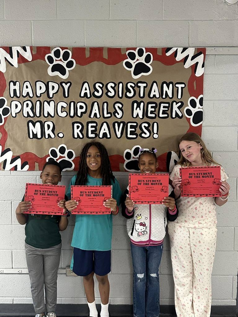 Four elementary students stand in front of a bulletin board that reads “Happy Assistant Principals Week Mr. Reaves!” Each student is smiling and holding a red “Bus Student of the Month” certificate. The display is decorated with paw prints and a red-and-black border on a school hallway wall.