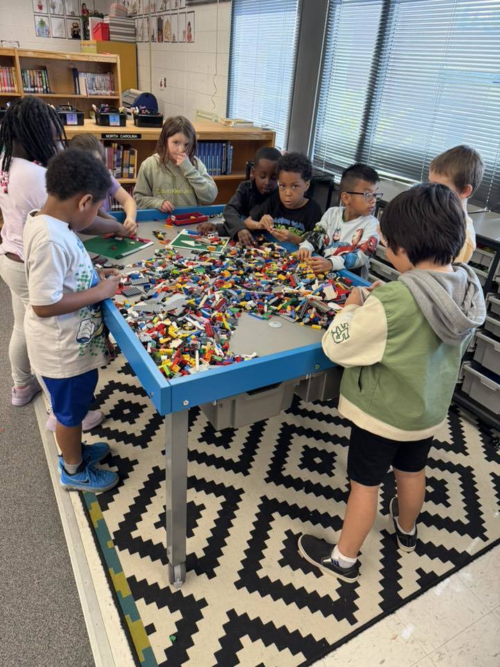 Eight elementary school-aged children gather around a blue activity table filled with a large quantity of colorful LEGO bricks in a classroom or library corner setting. The children appear engaged in playing with and building from the LEGOs, searching for pieces among the collection. The table features integrated grey bins underneath for storage.  The setting includes wooden bookshelves stocked with books in the background, with one shelf displaying a visible label reading "NORTH CAROLINA." The right wall features large windows covered with horizontal blinds. The floor beneath the table is covered by a decorative rug with a black geometric pattern on a white background, featuring a green/yellow border on one side and transitioning to grey carpet and possibly tile.
