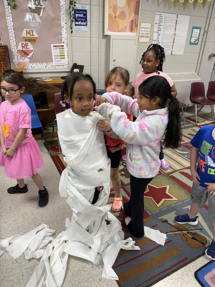 A diverse group of young children engage in a classroom mummy-wrapping activity. A Black girl with braided hair stands as the "mummy" being wrapped from the waist down with toilet paper by an Asian girl. A White girl watches from behind the mummy. A Black girl in a pink shirt observes with a smile further back. On the left, a White girl with glasses wearing a pink t-shirt and skirt watches the activity. A boy in a blue t-shirt marked "THE BEST" is partially visible on the far right.  The classroom features light-colored walls decorated with educational posters and artwork. Hexagonal shapes stacked vertically on the wall display musical notes: "SOL," "FA," "MI," "RE," "DO." A blue sign reads "You can't be great... if you can't audiate!" A yellow sign displays "You only get one chance to make a FIRST IMPRESSION." A large chart or grid labeled "Goal:" appears to track progress, possibly for "Grade 1," with rows marked by small stickers or indicators.  The floor is carpeted with a colorful design featuring a red star and a brown violin. Furniture including chairs and shelving are visible in the background.