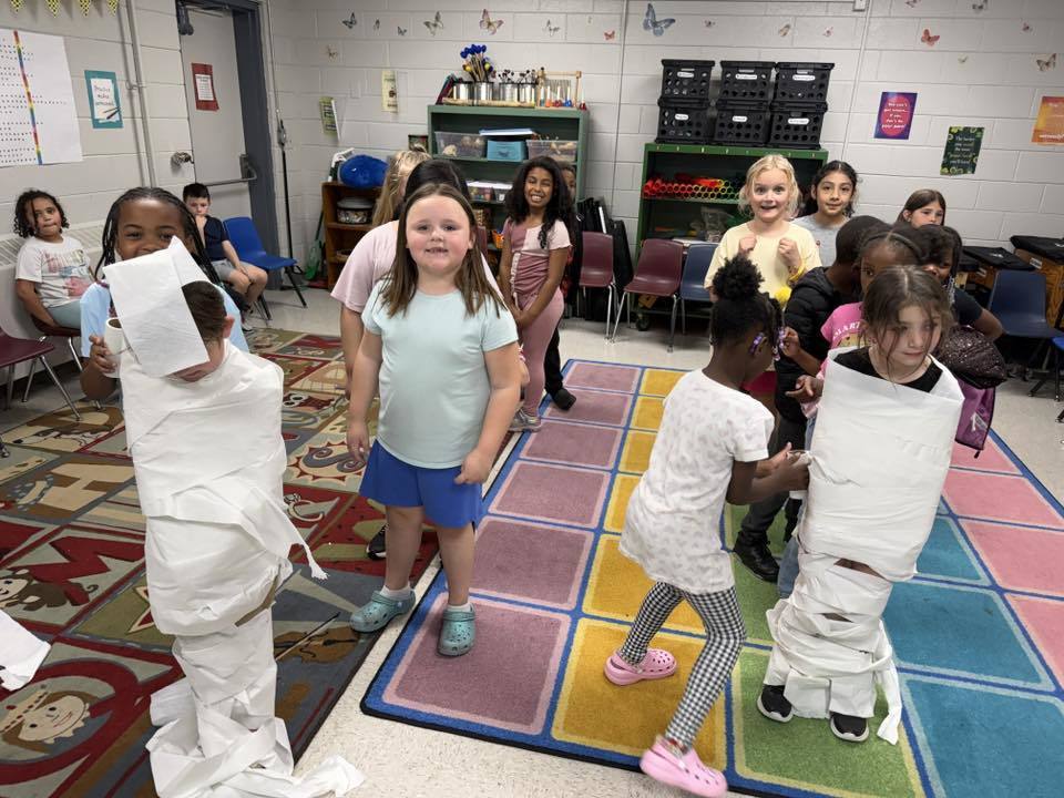  An elementary school classroom hosts a mummy-wrapping activity where children participate in wrapping their peers in toilet paper. Two children in the foreground are being wrapped to resemble mummies while their peers observe from various positions around the room. A girl in a light blue shirt stands prominently in the center, smiling at the camera.