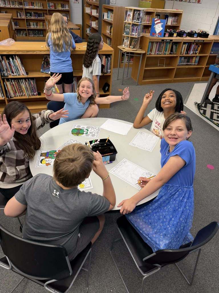 A group of elementary school-aged children engage in library or classroom activities. Six children gather around a light-colored round table in the foreground, with two girls and one boy looking at the camera and smiling, some waving. Papers with coloring activities, pencils, and markers are spread on the table. A black container labeled "TABLE 7" holds writing utensils.