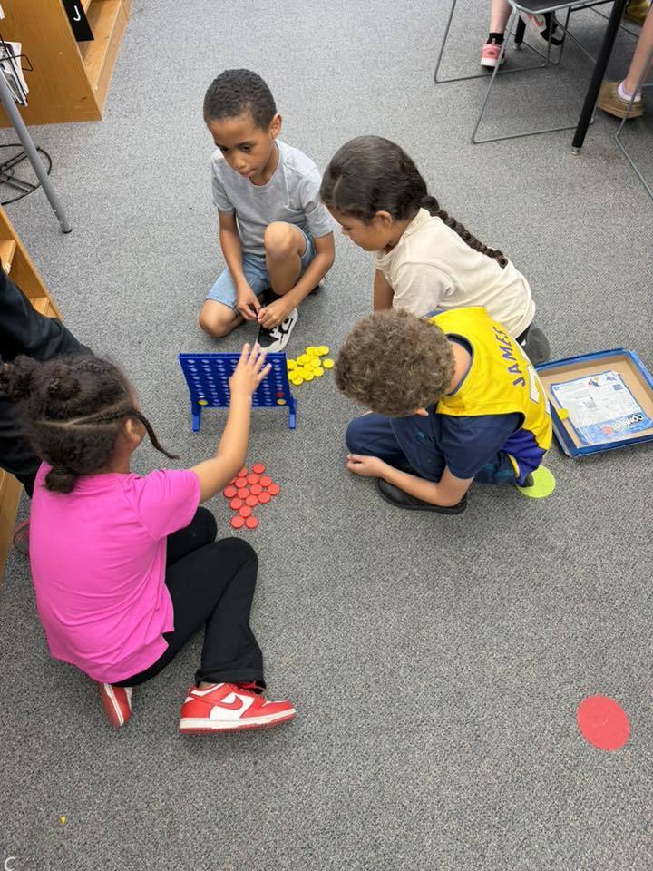Four elementary school-aged children sit on a grey carpeted floor playing Connect Four in a high-angle photograph. A blue Connect Four game board is positioned in the center with piles of yellow and red game pieces nearby.  From the top-left, a boy in a light grey t-shirt and blue shorts kneels while adjusting his white and black sneakers, looking down at the game. To his right, a girl with dark braided hair wearing a light-colored t-shirt focuses on the game. To the right of the board, a boy with curly hair wearing a yellow t-shirt with "JAMES" written on the back crouches while studying the board. On the left side, a girl in a bright pink t-shirt and black pants with red and white sneakers reaches her hand toward the game board to make a move.  In the background, a wooden shelving unit is visible on the left with a letter "J" label on one shelf. A Connect Four game box sits on the floor to the right of the children. A red circular marker is visible on the carpet in the lower right portion of the image.