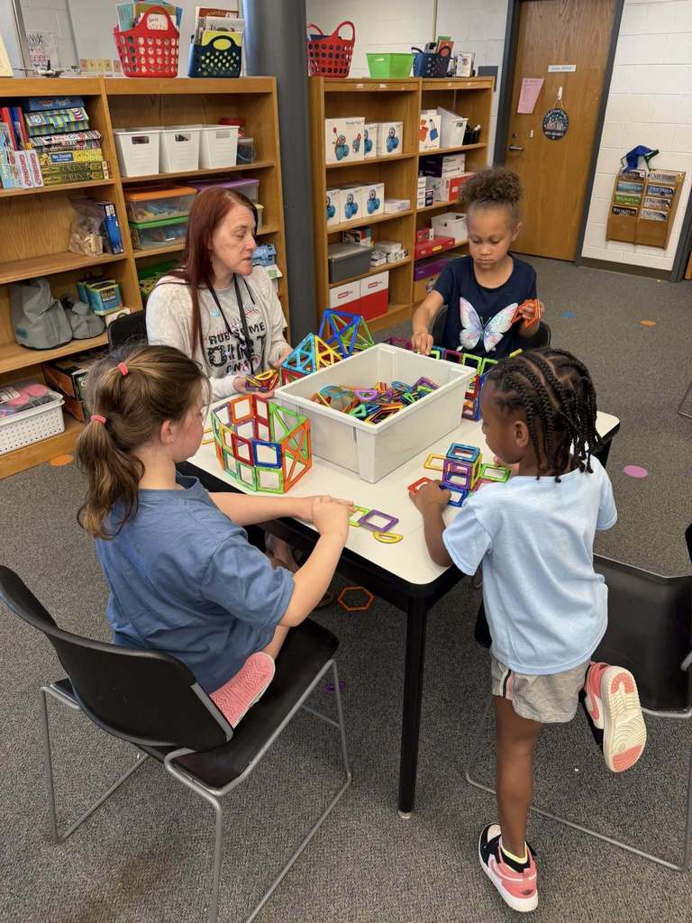 An adult woman and three young girls engage in a building activity with colorful magnetic tiles around a white rectangular table in a classroom or library setting. Two girls are seated on black chairs while another girl stands or kneels beside a chair. The adult woman, wearing a shirt printed with "AWESOME ORIGINAL," is also seated and participating in the activity. Colorful magnetic tiles are spread across the table and contained in a large white bin.  In the background, wooden bookshelves filled with books, baskets, and educational materials are visible. Boxes labeled "Wonder Pack" sit on the shelves. A brown door on the right displays a "STAFF LOUNGE" sign. Wall pockets next to the door are labeled "Our News" and "Our Why." The floor is carpeted with colorful circular markers.