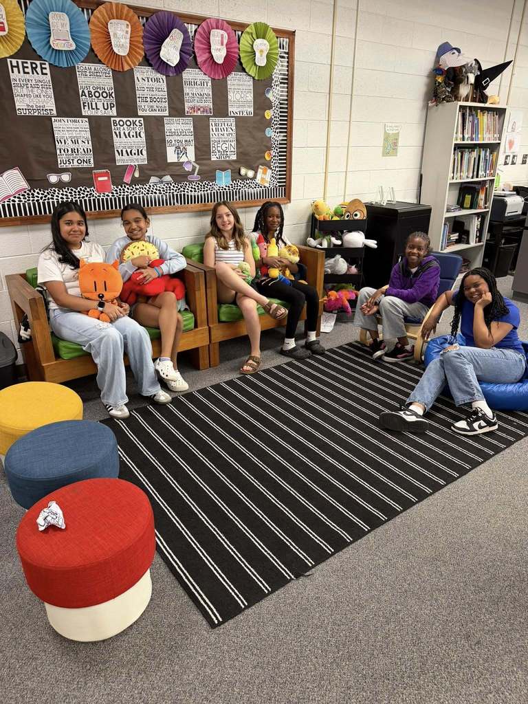 Six young girls gather in a school library or reading room, smiling at the camera. Four girls are seated in wooden chairs with green cushions, one sits in a blue beanbag chair, and another sits in a blue standard chair. Several girls hold colorful stuffed animals, with additional plush toys displayed on a cart in the background. Colorful round ottomans sit in the foreground, and a black rug with white stripes covers part of the grey carpeted floor.