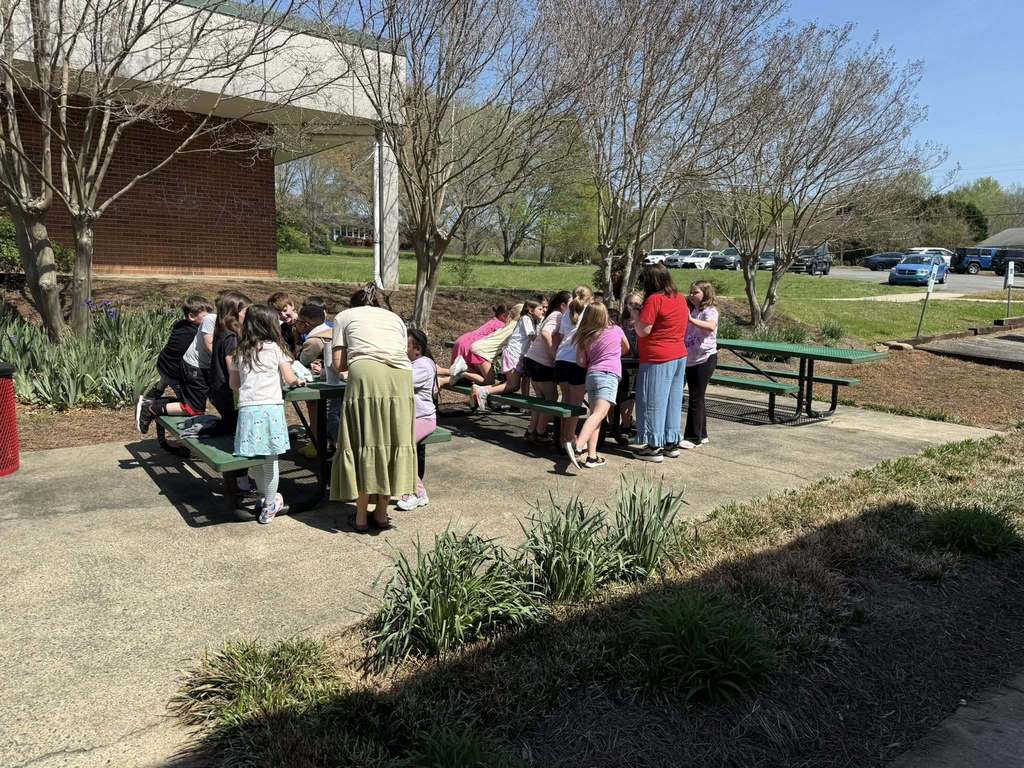 A group of children and several adults gather around two green picnic tables outdoors on a sunny day. The scene is set at a school with a brick building visible on the left and a parking lot containing several cars on the right. Bare-branched trees are scattered throughout the background, indicating late winter or early spring season.