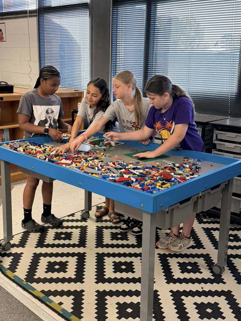 Four young girls gather around a blue rectangular table filled with a large collection of colorful LEGO bricks in an educational or creative classroom setting. The table features a recessed center area filled with bricks and a flat grey surface on the edges where girls place their creations or sort bricks. All four girls are actively engaged, looking down at the table and manipulating the bricks with their hands.
