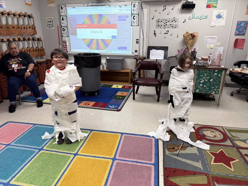 Two young children dressed as mummies, wrapped in toilet paper, stand on colorful rugs in a brightly decorated classroom. An adult woman sits on the left observing the children. The classroom features extensive musical and educational elements including a wall rack filled with ukuleles and musical notes drawn on the wall.