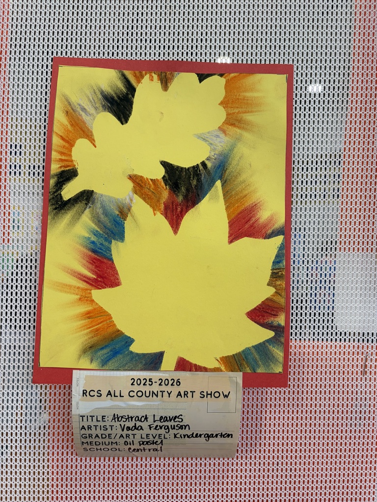 A kindergarten student artwork titled "Abstract Leaves" by Vada Ferguson is displayed as part of the 2025-2026 RCS All County Art Show from Central Elementary School. Created with oil pastel, the artwork features three yellow leaf silhouettes positioned against a background of radiating brushstrokes in red, orange, blue, and black, creating a dynamic burst effect. The artwork is framed with a red border and displayed on a white and red grid-patterned surface. A small white information card below the artwork provides details about the piece and its creator.
