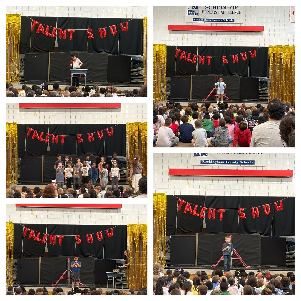 Students perform a variety of acts during a school talent show in the gym, including singing into microphones, speaking at a table, and performing as a group on stage in front of a large audience of seated students. A red “Talent Show” balloon banner hangs against a black backdrop with gold streamers on each side.