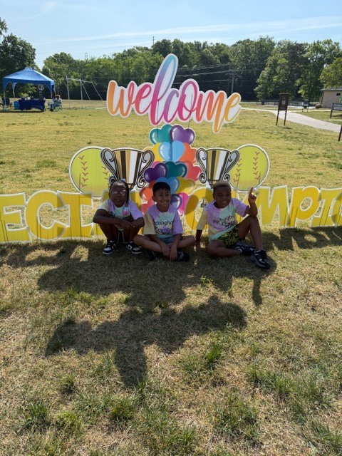 Three young children, likely boys, sit on a sunny grassy field in front of a large decorative sign celebrating achievement. The sign features "welcome" in colorful script at the top, followed by golden trophy cutouts, a yellow softball with red stitching on the left, and a cluster of colorful heart-shaped balloons in the center. Large yellow letters below spell "ECT" on the left and "MPTON" on the right, forming "CHAMPION" or "CHAMPIONS." The children wear tie-dye t-shirts and shorts, smiling at the camera with their shadows prominently cast on the grass in front of them. In the background, a wide grassy area, a blue canopy tent with people underneath, trees, power lines, and a paved path with a small building further back create the event setting.