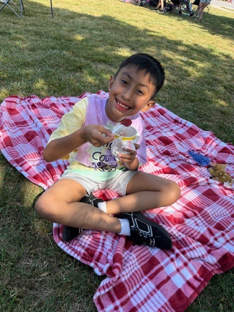 A young boy sits cross-legged on a red and white plaid blanket spread on green grass, smiling broadly at the camera. He holds a white cup with a yellow bottom in his left hand and a spoon with a red Kona Ice  in his right hand, appearing ready to eat it. Red residue is visible around his mouth. He wears a multi-colored tie-dye t-shirt in yellow, purple, and light green with faint, unreadable text, light-colored shorts, white socks, and black sneakers with white soles. On the blanket next to him are a small blue fuzzy object and a small light-colored stuffed animal. In the background, more green grass and blurry figures of people and outdoor equipment suggest an outdoor gathering or picnic. The scene is brightly lit by daylight.