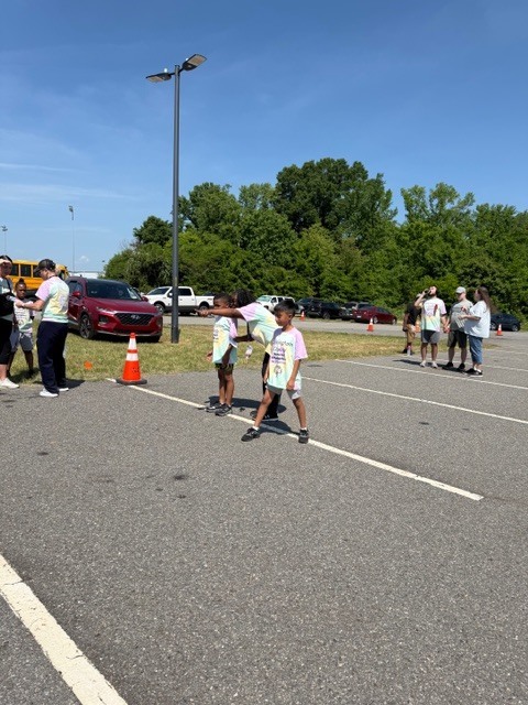 An outdoor event scene on a bright, sunny day captures two children in the foreground wearing colorful tie-dye shirts standing on an asphalt surface marked with white lines. The child on the right wears a tie-dye shirt with visible text reading "HAPPINESS" and additional text below. One child points toward the right. Further back on the right, a group of adults gathered together also wear tie-dye shirts. The background shows a parking area with several parked vehicles including a red SUV, a white pickup truck, and a yellow school bus partially visible on the far left. A tall black street light pole stands prominently on the left. Beyond the parking area, a dense line of green trees is visible under a clear blue sky. Orange traffic cones are positioned on the ground near the parked cars.