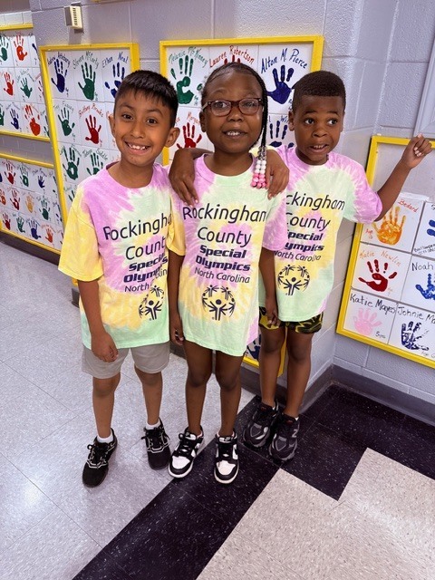 Three young children stand in a hallway smiling at the camera, all wearing matching tie-dye t-shirts printed with "Rockingham County Special Olympics North Carolina." The girl in the middle wearing glasses has her arms around the other two children. The boy on the left wears light-colored shorts and black sneakers. The girl in the middle wears a longer version of the t-shirt with black and white sneakers. The boy on the right wears patterned shorts and black sneakers with his left hand raised.
