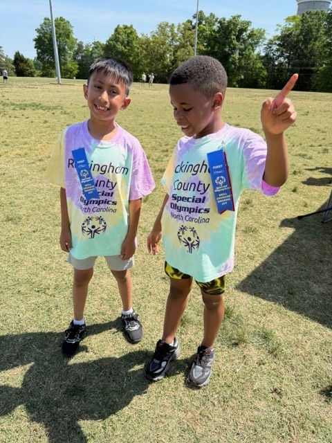 Two young boys stand on a sunny, grassy field outdoors wearing matching tie-dye t-shirts displaying "Rockingham County Special Olympics North Carolina" with the Special Olympics logo. Each boy wears a blue ribbon pinned to his shirt reading "FIRST SPECIAL OLYMPICS YOUNG ATHLETES SKILLS SHARING JOY," indicating achievement or participation recognition. The boy on the left smiles and looks at the camera, while the boy on the right looks slightly away and upward, pointing his right index finger toward the sky. In the background, trees, distant figures, and a tall pole suggest a park or athletic field setting.