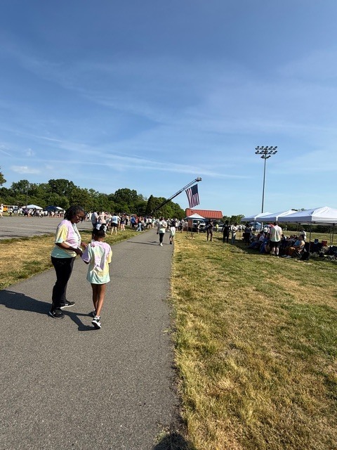A wide-angle photograph captures an outdoor community event under a clear blue sky. In the foreground, an adult and a child, both wearing tie-dye shirts, walk away from the camera on a paved path. To their right is a large expanse of dry, yellowish grass. Further down the path, more people are visible walking and gathering. On the right side of the image, several white canopy tents are set up with groups of people congregated around them, some sitting. In the midground, an American flag is suspended from an extended boom or ladder of what appears to be a fire truck. A tall light pole with multiple floodlights stands further to the right. A line of green trees marks the horizon in the background, creating a scenic outdoor setting for this community gathering.
