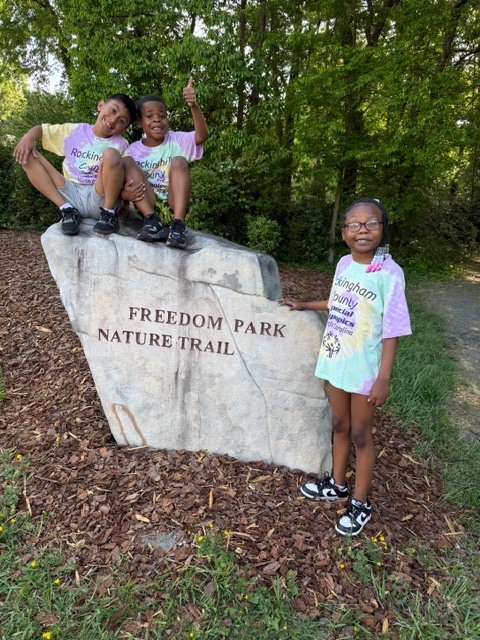  Three children pose next to a large stone sign reading "FREEDOM PARK NATURE TRAIL" in a park setting. Two boys sit on top of the stone sign, with one giving a thumbs-up gesture. A girl stands to the right of the sign, resting her hand on it. All three children wear matching tie-dye t-shirts printed with "Rockingham County Special Olympics North Carolina." The background features trees and greenery, with wood chips covering the ground around the sign. The children's cheerful poses and matching apparel indicate participation in a Special Olympics event or celebration.