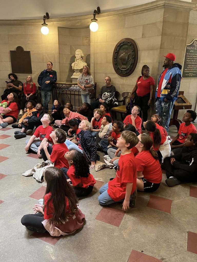 A group of elementary school students wearing matching red T-shirts sit on the floor inside a historic-looking building with stone walls and plaques. They look upward attentively, as if listening to a guide. Several adults stand behind them, and a white bust sculpture and wall-mounted displays are visible in the background.