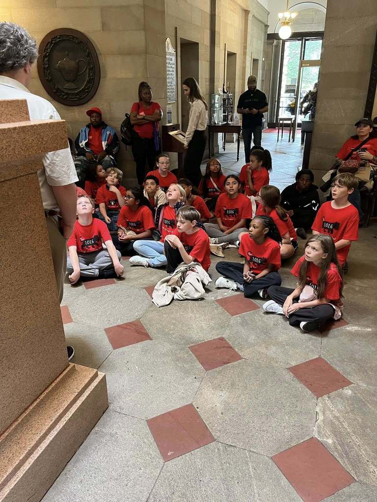 "A large group of elementary school children wearing red t-shirts labeled '1016 Our History' sit on the stone floor of a museum hall with hexagonal tile patterns, listening attentively to an adult educator. Stone walls, ornate ceiling fixtures, and architectural details are visible in the background, with several adult chaperones standing nearby."
