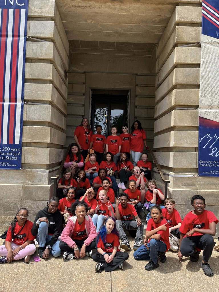 "Approximately 30-40 children and adult chaperones, mostly wearing red t-shirts, pose together on the classical stone steps of a historic building. Banners on either side commemorate the 250-year founding anniversary of the United States, with the NC Department of Natural and Cultural Resources logo visible."