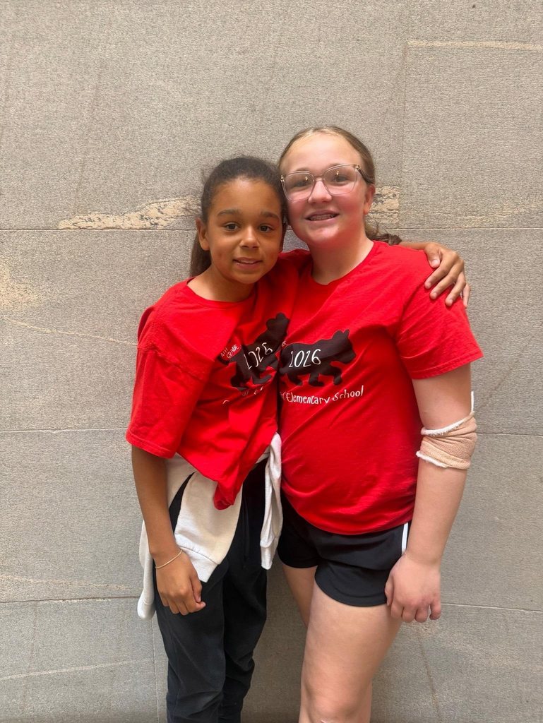 Two elementary school girls wearing matching red school T-shirts stand side by side against a stone wall, smiling at the camera. One girl has her arm around the other, showing a friendly, supportive pose.