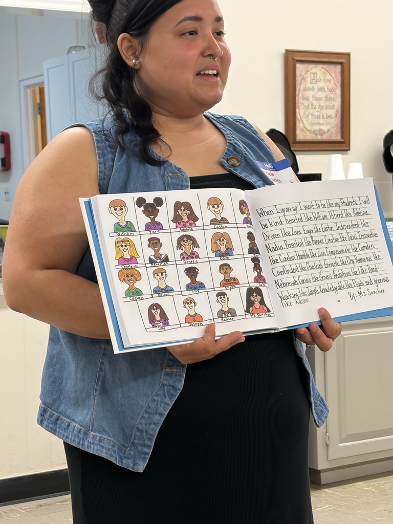 A woman with dark hair tied up wearing a denim vest over a black top holds an open book and smiles while looking toward the right. The book displays student artwork and a heartfelt message. The left page features cartoon-style drawings of children's faces arranged in rows, each labeled with names including William, Zara, Adelina, Easton, Charleigh, Nehemiah, Genesis, Cam, Caelan, Kaiser, Chy, Elijah, Lilke, Joseph, Banner, and others. The right page contains a handwritten paragraph by Ms. Sanchez that reads: "When I grow up I want to be like my Students. I will be kind-hearted like William. Patient like Adelina. Driven like Zara. Eager like Easton. Independent like Nadia. Persistent like Banner. Creative like Jani. Innovative like Caelan. Humble like Zion. Compassionate like Camden. Confident like Charleigh. Energetic like Chy. Humorous like Nehemiah. Curious like Genesis. Ambitious like Lillie. Hard-Working like Joseph. Knowledgeable like Elijah. and generous like Kaiser."  In the background, a framed picture on a light-colored wall displays religious text reading: "And now abideth faith, hope, love, these three; but the greatest of these is love. 1 Corinthians 13:13."