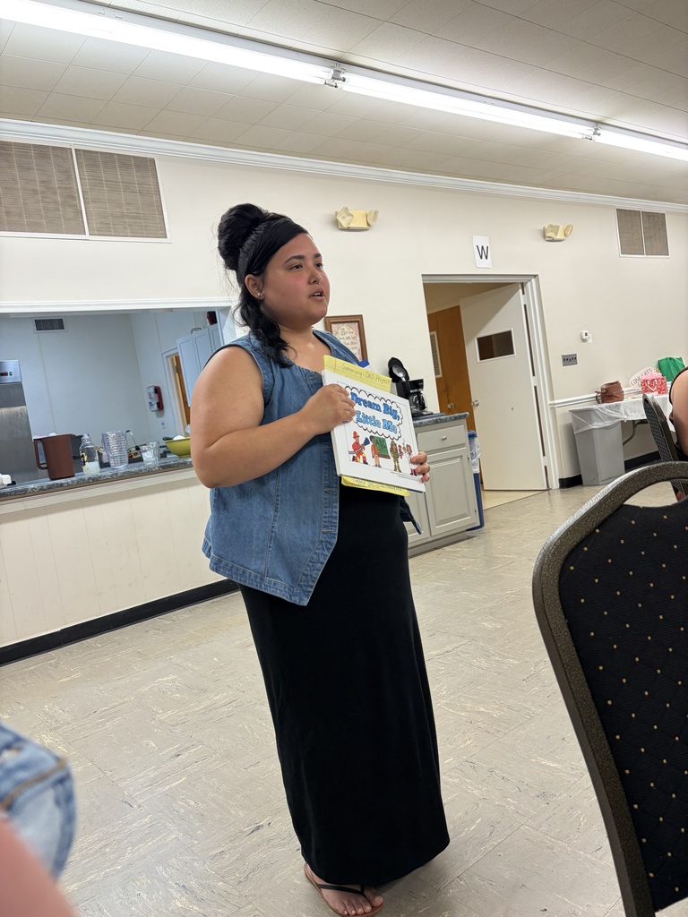 A woman with dark hair styled in a bun with a headband stands indoors holding a children's book titled "Dream Big, Little Me." She wears a denim vest over a black long dress or skirt with flip-flops and appears to be addressing an unseen audience with her mouth slightly open. The setting is an indoor room with light-colored walls and floor tiles, suggesting a community center or multi-purpose room. A counter area with kitchen items is visible on the left. On the right, a doorway marked with the letter "W" is visible along with chairs and a trash bin. Fluorescent lighting fixtures are mounted on the ceiling, and an air vent is visible on the upper left wall. A dark brown chair with a gold patterned seat is partially visible in the bottom right corner.