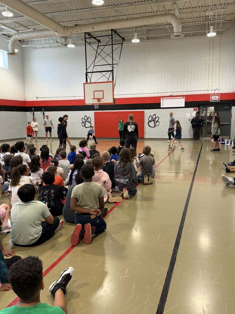 "A large group of elementary school students sit on the gymnasium floor in rows, watching adults and older children who are standing in the center participating in a game or activity. The gym features light-colored flooring with red and black markings, white walls with red padding, large black paw print decals, a basketball hoop with backboard, and a green EXIT sign on the far right wall."