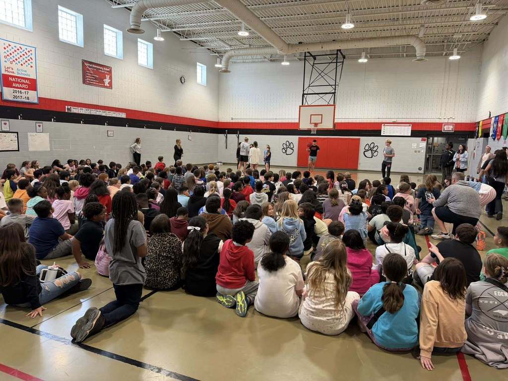 "Elementary school students sit on the gymnasium floor facing the front wall. Adults and children stand at the front near a basketball hoop. One person holds a basketball. The gym has white cinder block walls with red and black horizontal stripes, a basketball hoop with red rim, and black paw print logos on the wall."