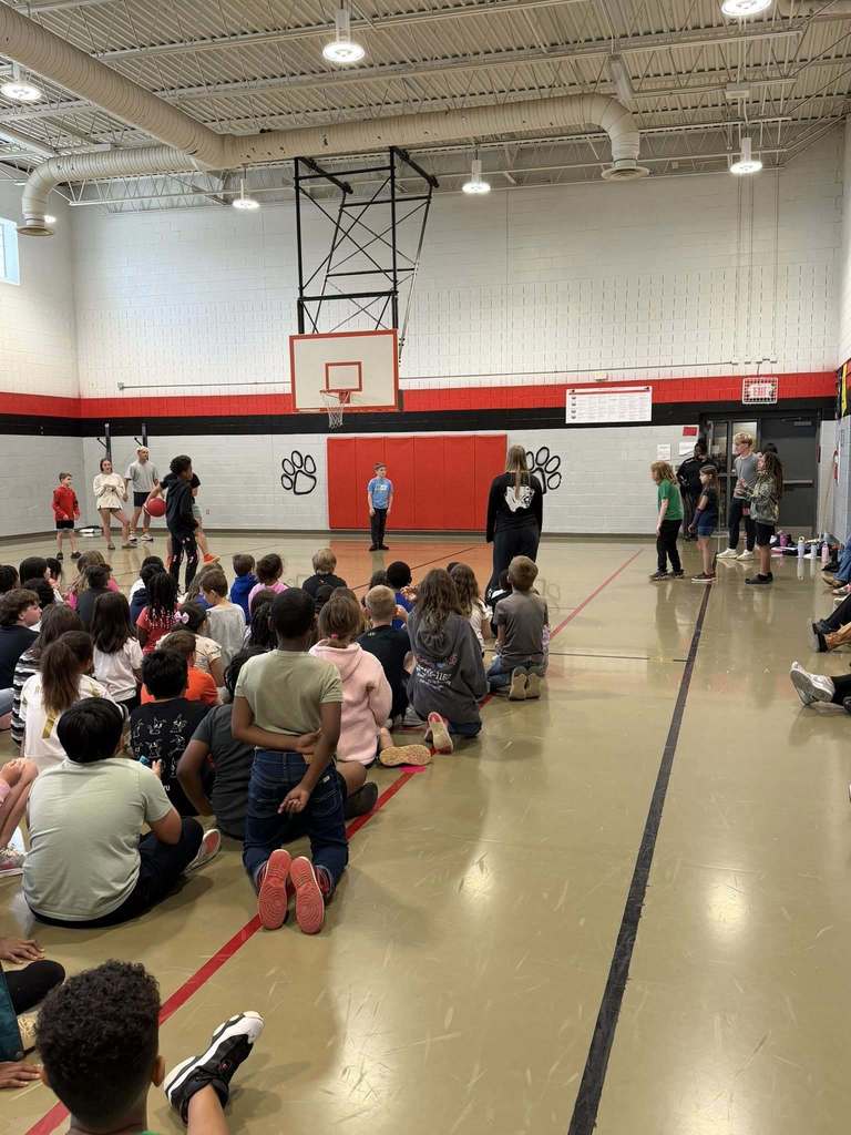 "A large group of elementary school students sit on the gymnasium floor facing adults and older students standing near the far wall. One older student holds a basketball. Banners on the left wall show National Awards for 2016-17 and 2017-18. The gym features gray and white cinder block walls with red and black stripes, a basketball hoop, flags from different countries on the right wall, and black paw print graphics on the red wall section."