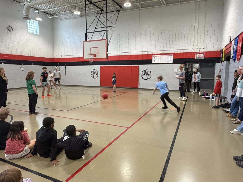"A boy in a light blue shirt runs toward a red kickball on a gymnasium floor during a kickball game. A girl stands near the ball facing him. Elementary school students sit on the floor watching the game, while adults and children stand along the walls observing. The gym has white brick walls with red and black horizontal stripes, a basketball hoop, black paw print logos on the back wall, and an EXIT sign visible on the right wall."