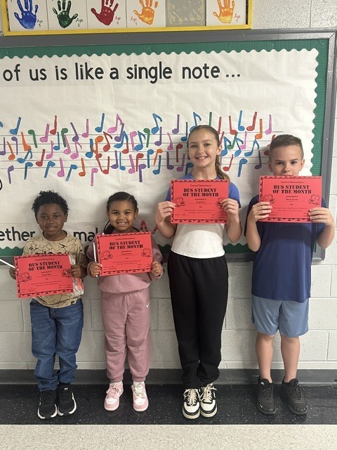 "Four smiling elementary school students stand in a line holding red 'Central Elementary's Bus Student of the Month' certificates. They are positioned in front of a colorful bulletin board decorated with musical notes and a banner featuring handprints labeled with ages 4, 4, 7, and 9. The bulletin board text reads 'of us is like a single note...'"