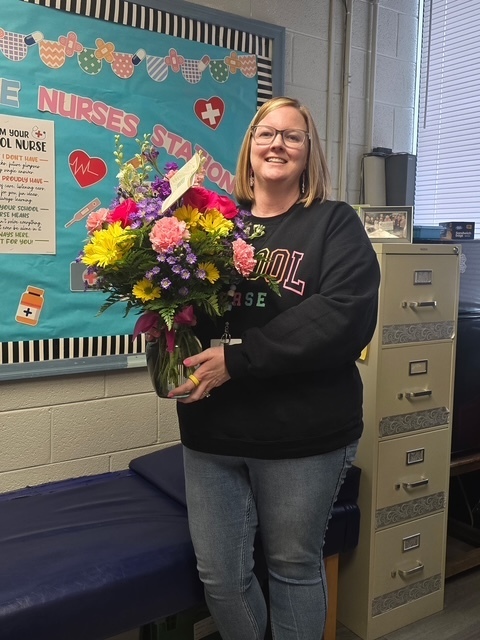  A smiling woman with blonde hair and glasses holds a large, colorful bouquet of flowers in a glass vase. She wears a black sweatshirt printed with "SCHOOL NURSE" in colorful letters and blue jeans, with a yellow ring on her right hand. Behind her is a light blue bulletin board adorned with healthcare-related cut-outs including hearts, pills, crosses, stethoscopes, and bandages along the top border