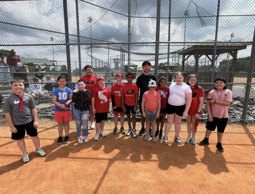 Thirteen individuals—primarily children with a few young adults—stand in a line on a reddish-brown dirt baseball field, facing the camera and mostly smiling. They wear a mix of sports attire, with several in red team shirts. Visible text on clothing includes "10," "1016 TRACK & FIELD," "2016" on a white baseball cap and shirt, "CENTRAL WILDCATS," "EDEN SOC," and "EDEN." A tall chain-link fence is visible behind them, with other parts of a sports complex including additional fences, a covered structure, and observing spectators visible in the background. The partly cloudy sky is visible above. Long shadows of the individuals are cast on the dirt in front of them, indicating the sun is positioned behind the group.