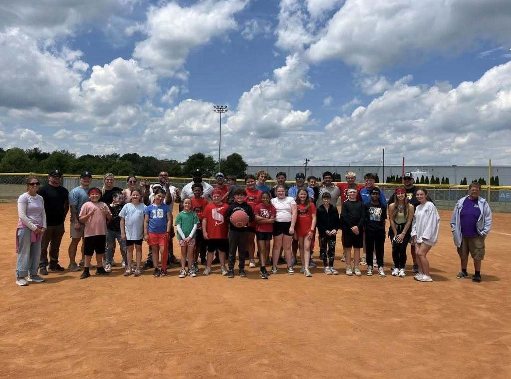 Approximately 30-35 people—consisting of adults and children/teenagers—pose for a photograph on a dirt baseball or softball field arranged in several rows. Adults are positioned primarily on the ends and in the back rows, while children and teenagers are arranged in the middle and front rows. Many individuals are smiling. One child in the center holds a basketball. The sky above is mostly blue with scattered white clouds. In the background, a yellow chain-link fence, a utility pole with stadium lights, trees, and industrial-looking buildings are visible, creating a sports complex setting.