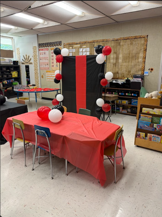 A classroom or multi-purpose room is set up for an event with festive red, black, and white party decorations. In the foreground, a rectangular table is covered with a red plastic tablecloth and decorated with red and white balloons. Four small chairs in yellow, blue, and green are positioned around the table. To the left, a blue table with a red top is set with clear plastic cups and red plates. A decorative backdrop made of black and red plastic sheeting stands in the middle background, flanked by balloon stands holding red, black, and white balloons.