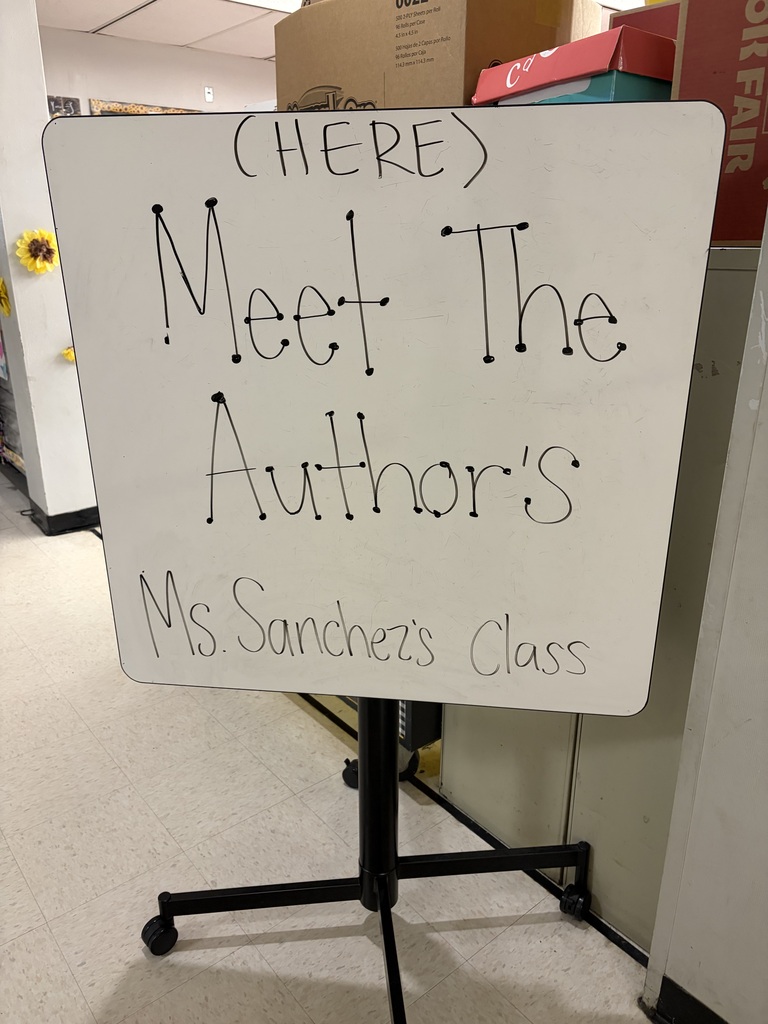 A white dry-erase board on a black wheeled stand displays handwritten text reading "Meet The Author's Ms. Sanchez's Class" with an arrow pointing to "(HERE)." The setting appears to be an indoor school or community space with tiled flooring and white walls. In the background, cardboard boxes are stacked, with one box displaying product information including "500 2-PLY sheets per Roll," "16 Rolls per Case," "4.5 ins x 4.5 ins," and Spanish text "500 hojas de 2 Capas por Rollo," "16 Rollos por Caja," and "114.3 mm x 114.3 mm." A yellow sunflower decoration is partially visible on the left side.