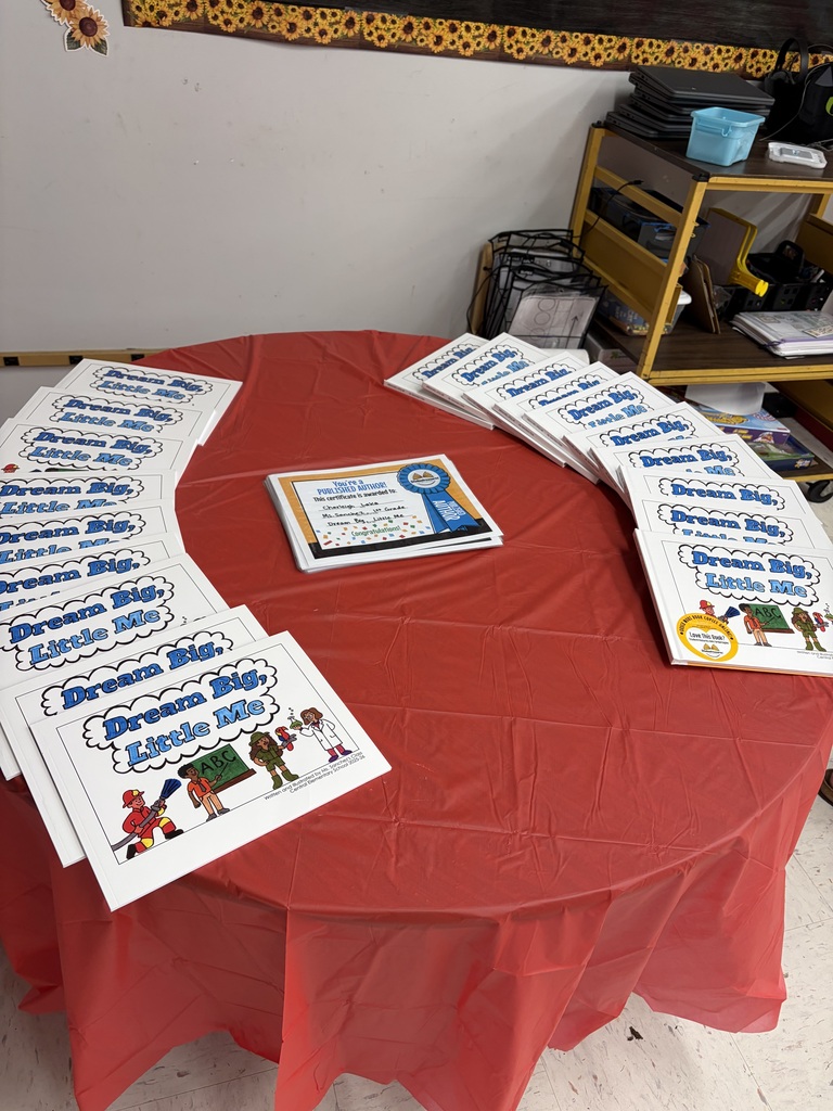 A round table covered with a red plastic tablecloth displays the published children's book "Dream Big, Little Me" arranged in a semi-circle. Each book cover features illustrations of four children dressed as various professionals: a firefighter, a person holding an "ABC" block, a soldier, and a scientist or doctor. One book cover displays a "Love This Book!" sticker. Small text on the book covers reads "Written and Illustrated by Ms. Spencer's Central Elementary School 2023-24." In the center of the table, a stack of certificates is visible, with the top certificate reading: "You're a PUBLISHED AUTHOR! This certificate is awarded to: Sharleigh, Iasa, Ms. Sanchez' 3rd Grade, Dream Big, Little Me - Congratulations!" The background shows a classroom or school setting with a whiteboard, a decorative sunflower border, and a shelf unit filled with various school supplies and electronics.