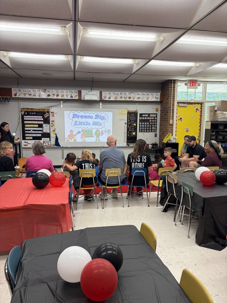 A classroom or multi-purpose room is set up for a student author event featuring a presentation of "Dream Big, Little Me." Several adults and young children are seated facing a whiteboard displaying the book title and the date "5-5-26." The room is decorated with standard classroom elements including an alphabet frieze displaying "Aa Bb Cc Dd Ee Ff Gg Hh Ii Jj Kk Ll Mm Nn Oo Pp Qq Rr Ss Tt Uu Vv Ww Xx Yy Zz," an "I CAN..." poster on the left wall with partially visible bullet points about creating, editing, writing, asking questions, and telling stories, a calendar showing April, and a clock displaying 1:09. An "EXIT" sign is visible on the wall, and a window is present in the room. In the foreground, tables are covered with red and black tablecloths and decorated with red, white, and black balloons, creating a festive atmosphere. Attendees are seated on small chairs with their backs mostly to the camera, focused on the whiteboard presentation. Some children are wearing t-shirts with text including "THIS IS HOME" and other partially visible text. The room features fluorescent lighting typical of a school setting.