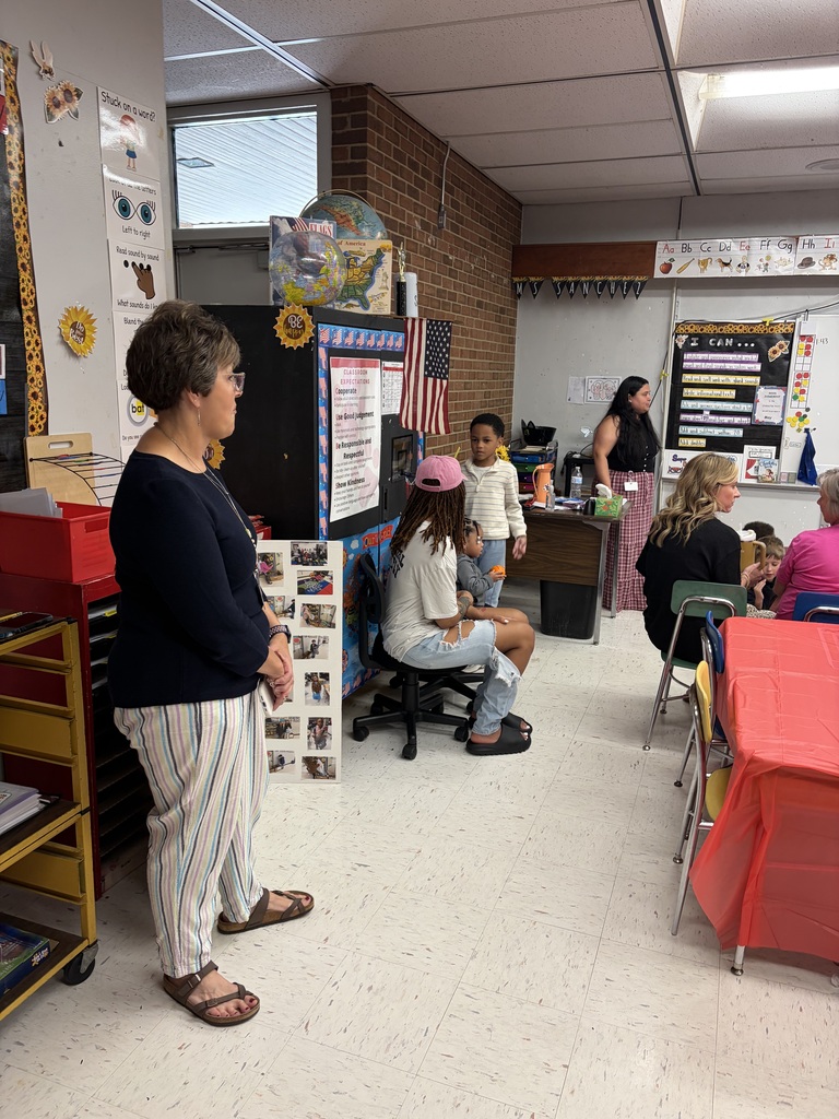 A lively classroom setting hosts an educational event with several adults and children engaged in various activities. On the left, an adult woman stands observing the scene. In the center, an adult wearing a pink cap sits on a chair with a young child on their lap while a boy stands beside them. To the right, another adult stands near a whiteboard, seemingly leading a discussion or activity, while multiple adults and children are seated at tables covered with red tablecloths.