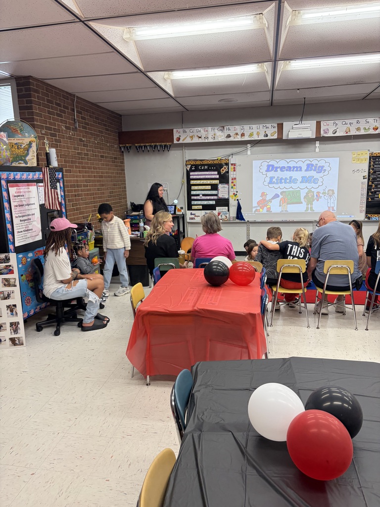 A classroom event celebrates the publication of "Dream Big, Little Me" with a book launch and author celebration. The classroom features educational displays including an alphabet line across the top wall reading "Aa Bb Cc Dd Ee Ff Gg Hh Ii Jj Kk Ll Mm Nn Oo Pp Qq Rr Ss Tt Uu Vv Ww Xx Yy Zz," a "CLASSROOM EXPECTATIONS" poster on the left wall listing "Cooperate, Use Good Judgement, Be Responsible and Respectful, Show Kindness," and an "I CAN..." board with learning objectives in reading, writing, math, social studies, and science. An American flag and globe are visible on shelves. A large projection screen displays "Dream Big, Little Me 5-15-26" with cartoon illustrations representing different professions: a firefighter, an "ABC" block, a person holding a clipboard, and a scientist with a beaker. Several adults and children are gathered in the room, with children seated on small chairs facing the screen. Two tables are set up in the foreground—one red with red and black balloons, and one black with white, black, and red balloons, creating a festive atmosphere. Standard classroom fluorescent lighting panels are visible on the ceiling.