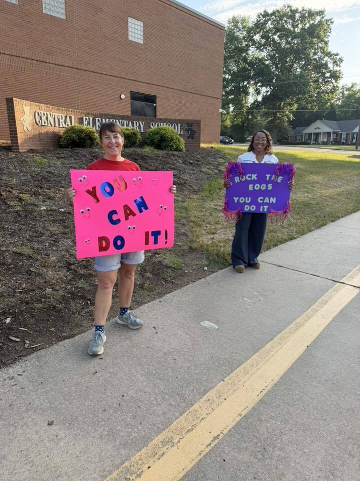 Two women stand on a sidewalk in front of Central Elementary School holding encouraging signs for students taking EOG tests. The woman on the left holds a bright pink sign with blue and pink letters reading "YOU CAN DO IT!" surrounded by small cartoon faces. The woman on the right holds a purple sign with fringe reading "ROCK THE EOGS YOU CAN DO IT." Both women are smiling. The background features trees, other buildings, and a bright sky in front of the brick school building.