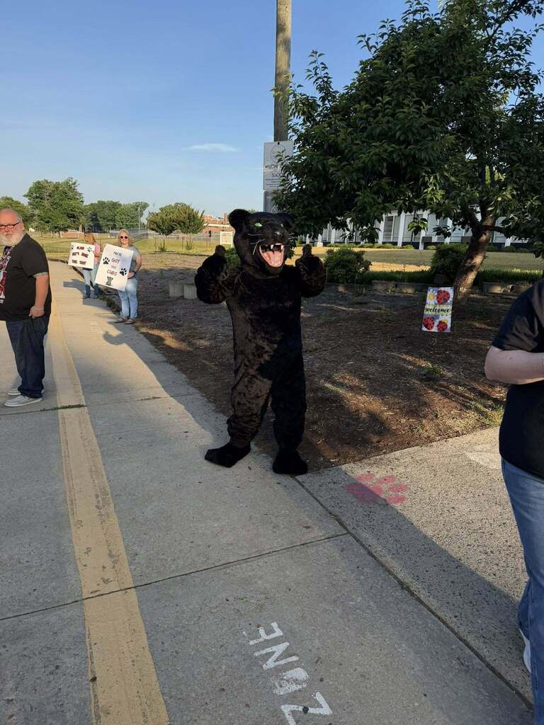 A black panther mascot stands on a sidewalk giving two thumbs up in a supportive gesture. Behind the mascot, two women hold signs with paw prints and messages: "WE LOVE OUR CUBS!" and "YOU'VE GOT THIS CUBS!" A man stands further left on the sidewalk. A "WELCOME" sign is visible next to a tree.